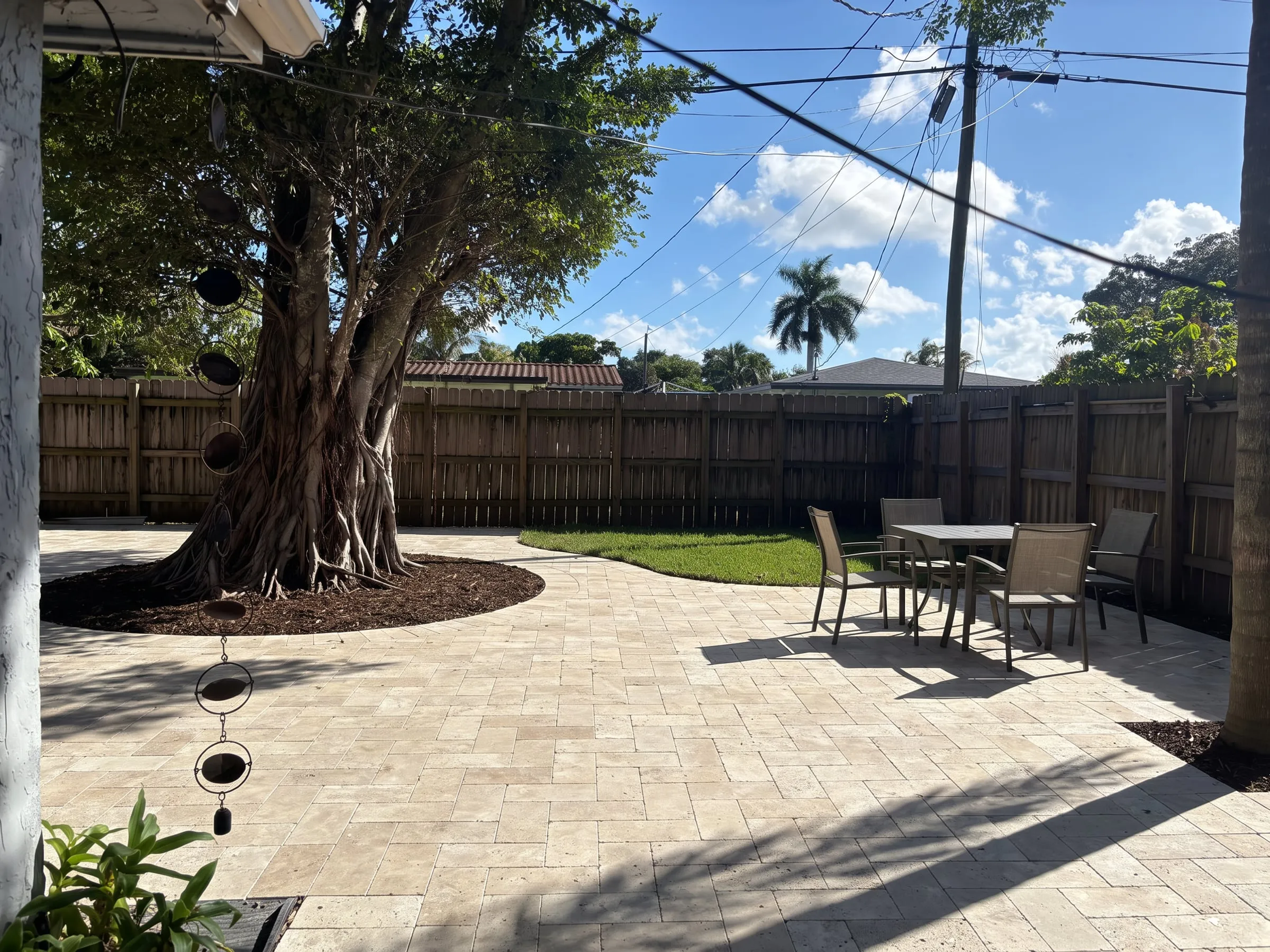 AI visualization of the same backyard as a travertine paver patio with a dining set and planted tree ring
